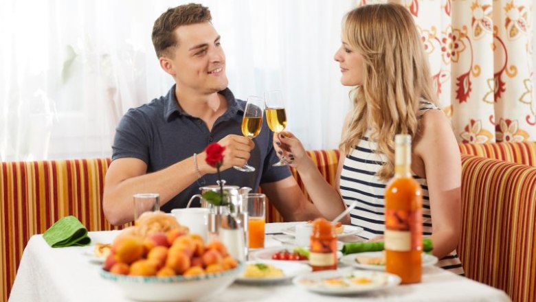 Ein Paar stößt mit Sektgläsern beim Frühstück an, umgeben von Obst und Getränken auf einem gedeckten Tisch.