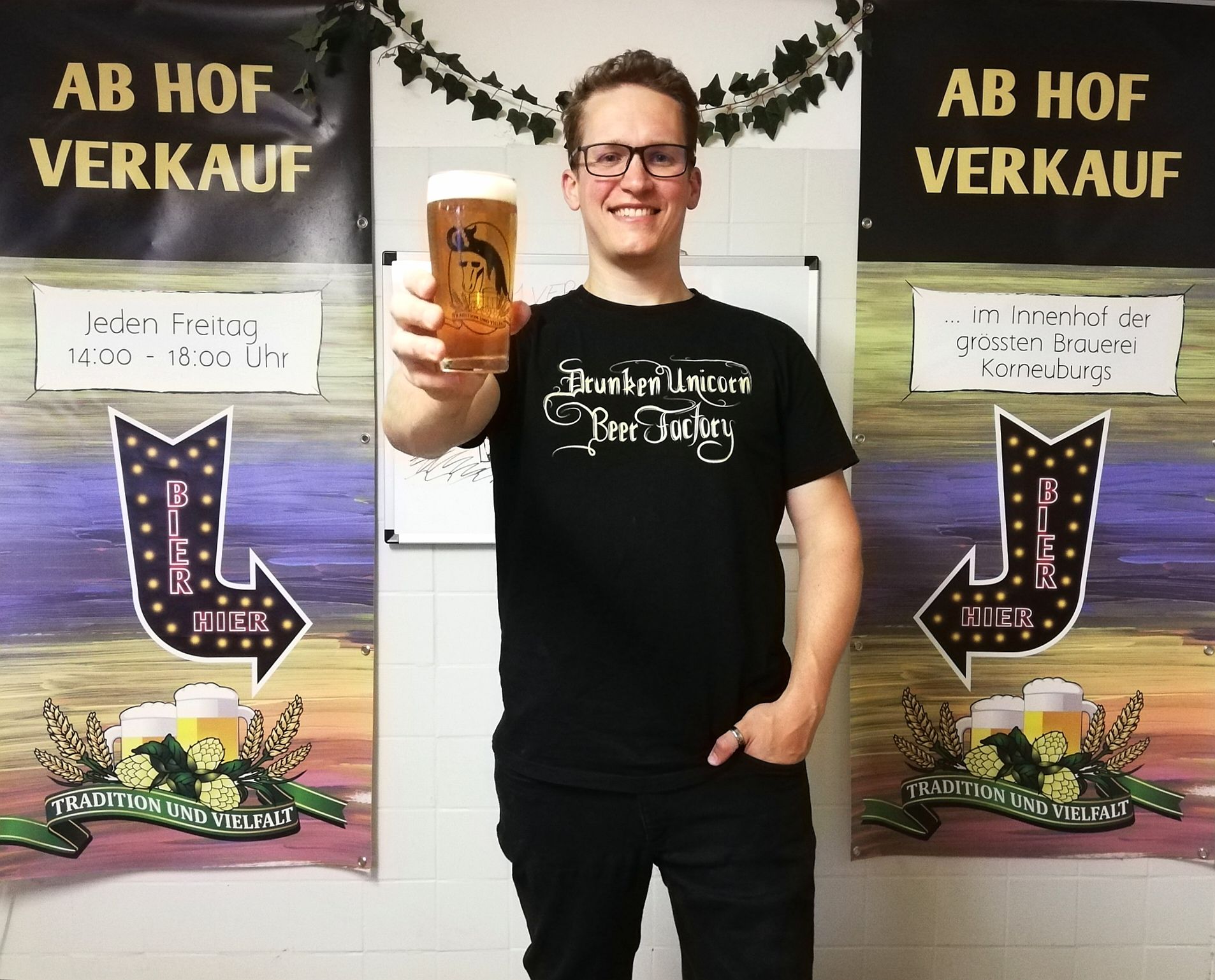 Person hält ein Bierglas vor zwei Bannern mit der Aufschrift 'Ab Hof Verkauf'.