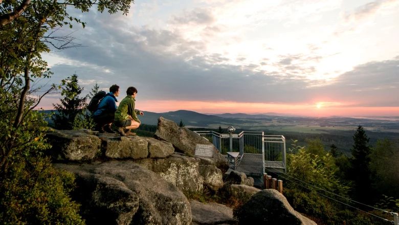 Zwei Personen auf einer Felsformation bei Sonnenuntergang mit Blick auf eine Aussichtsplattform und weite Landschaft.