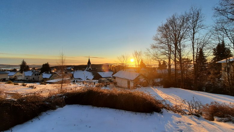 Winterlandschaft mit Sonnenaufgang &uuml;ber einem verschneiten Dorf und kahlen B&auml;umen.