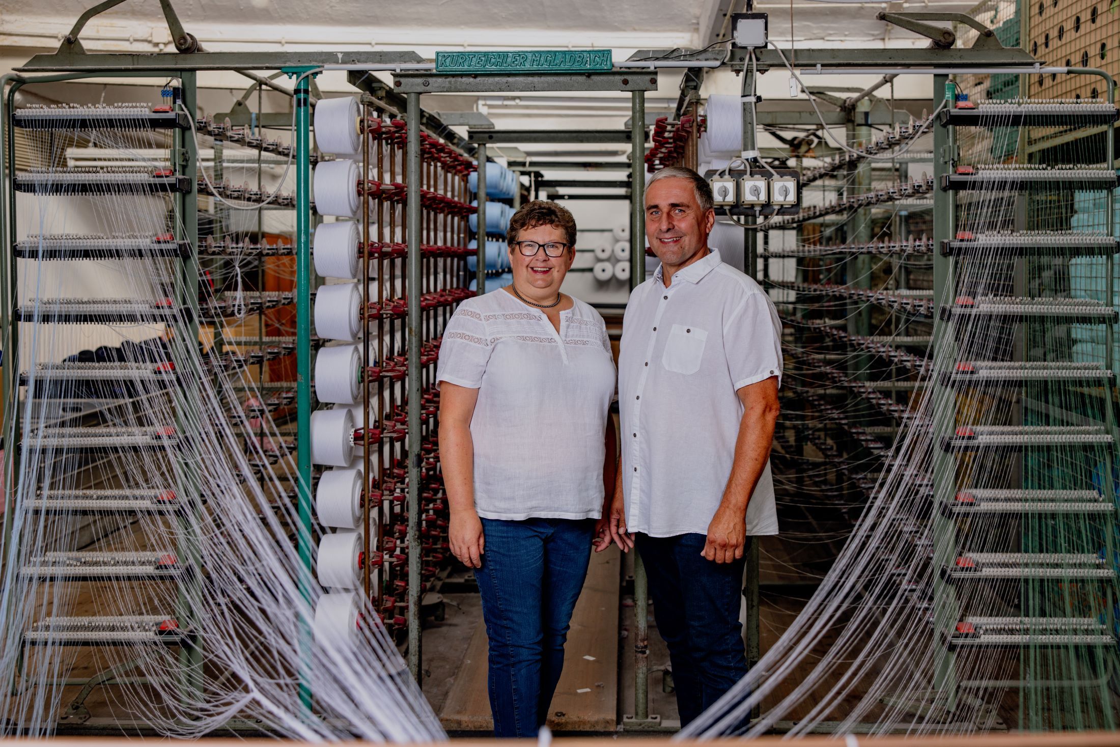 Zwei Personen stehen in einer Textilfabrik vor Maschinen mit Garnrollen.