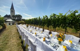 Lange, festlich gedeckte Tafel im Freien neben einem Weinberg, im Hintergrund eine Kirche.