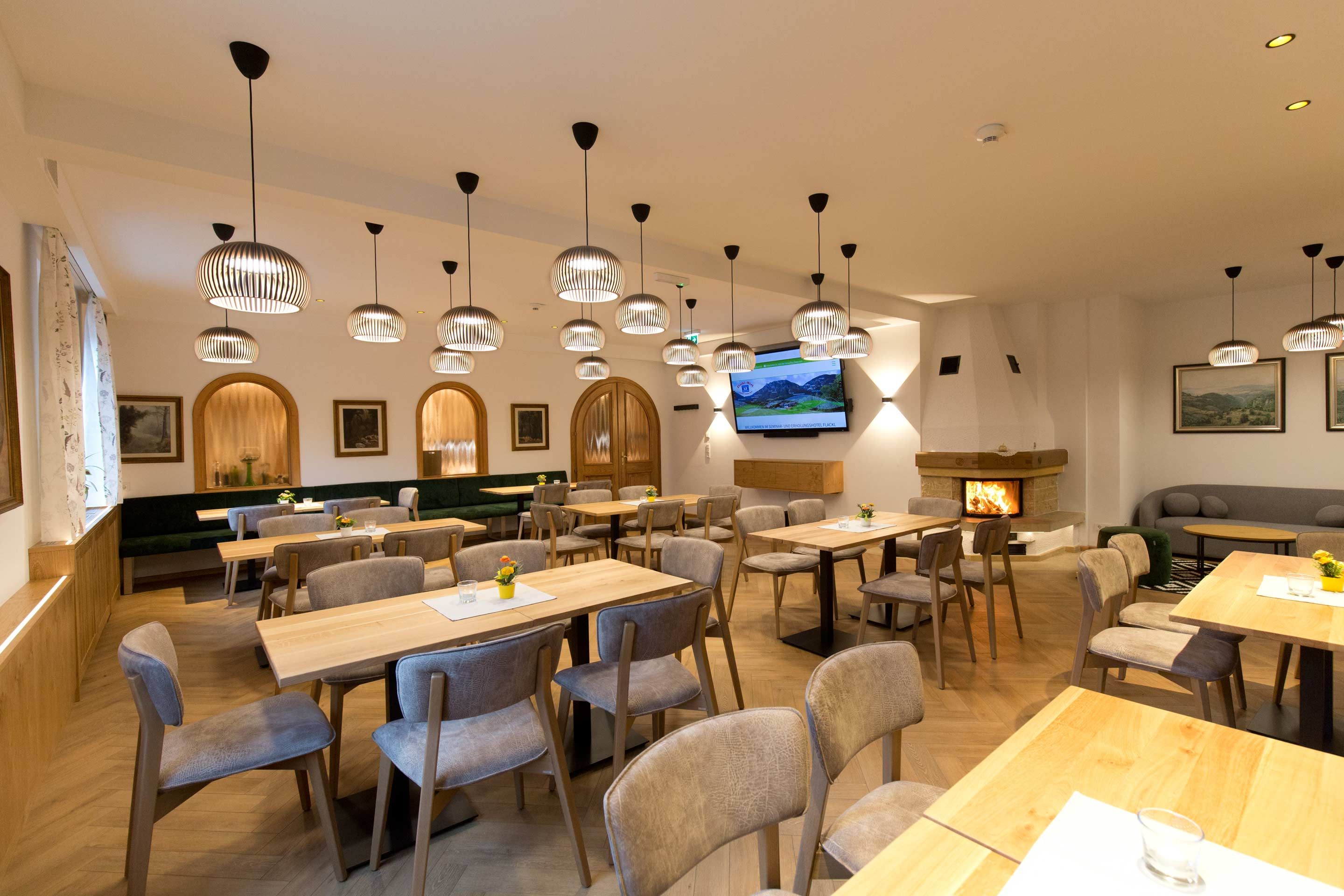 Ein gemütliches Kaminzimmer mit Holztischen, Stühlen und modernen Lampen. Ein Kamin brennt im Hintergrund.