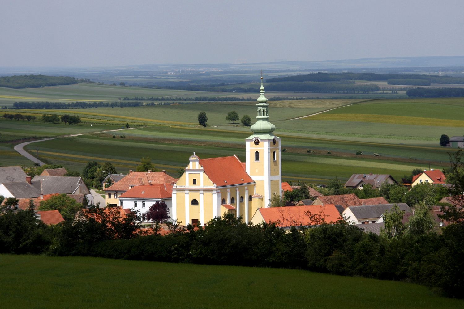 Blick auf eine Kirche in Ottenthal mit umliegenden Häusern und weiten Feldern im Hintergrund.