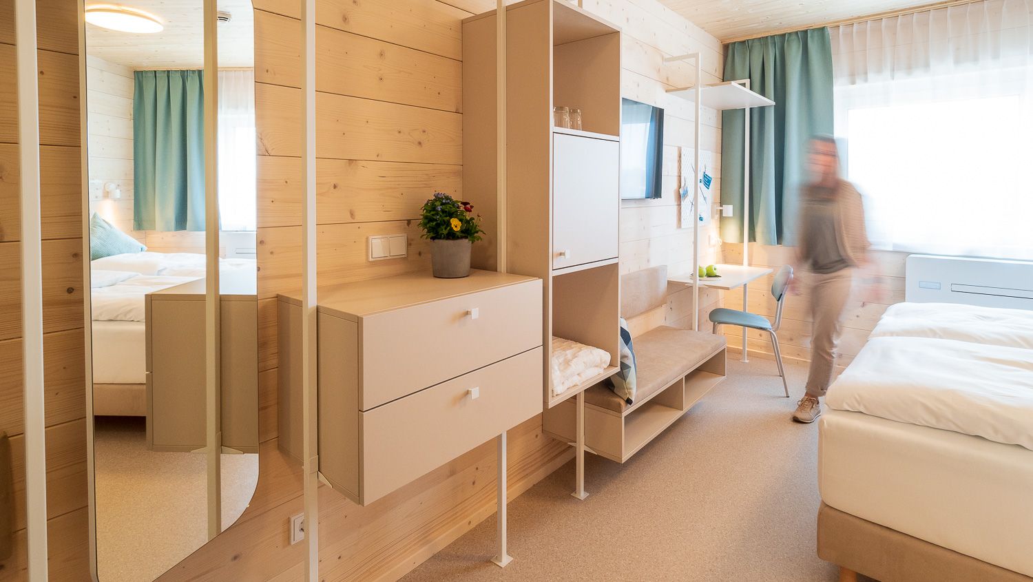 Modernes Hotelzimmer mit Holzverkleidung, Spiegel, Schränken und einem Bett. Eine Person bewegt sich im Raum.