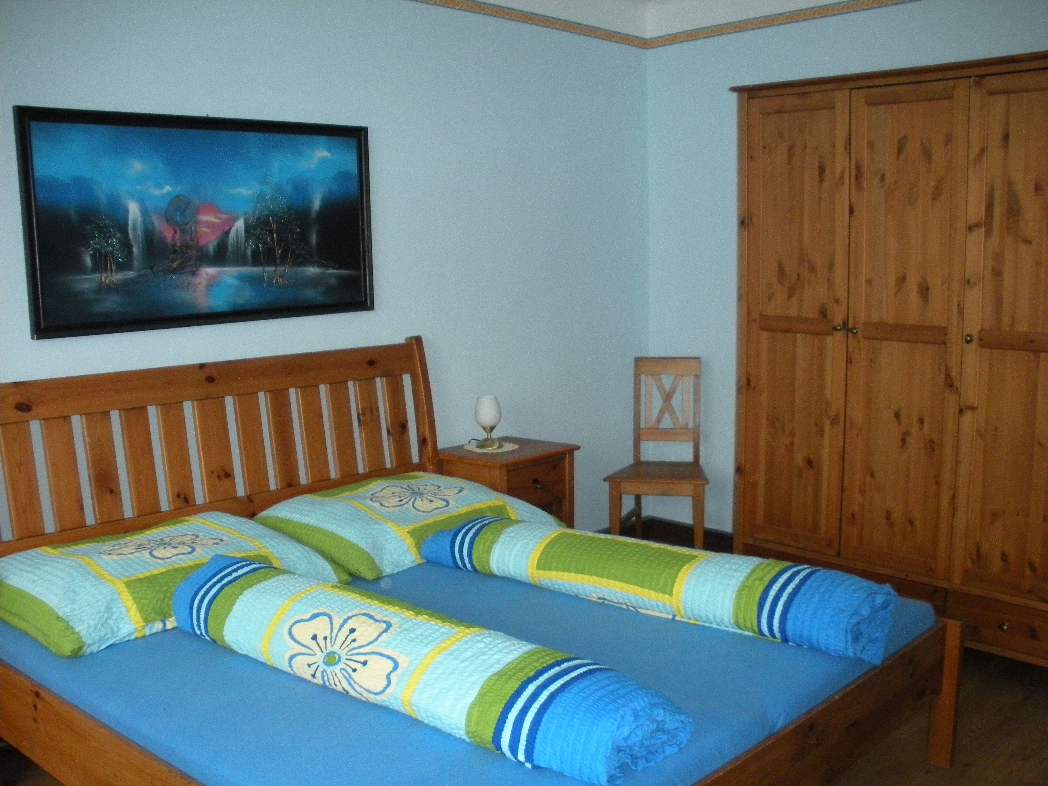 Ein Gästezimmer mit einem Holzbett, bunten Bettdecken, einem Schrank und einem Bild an der Wand.