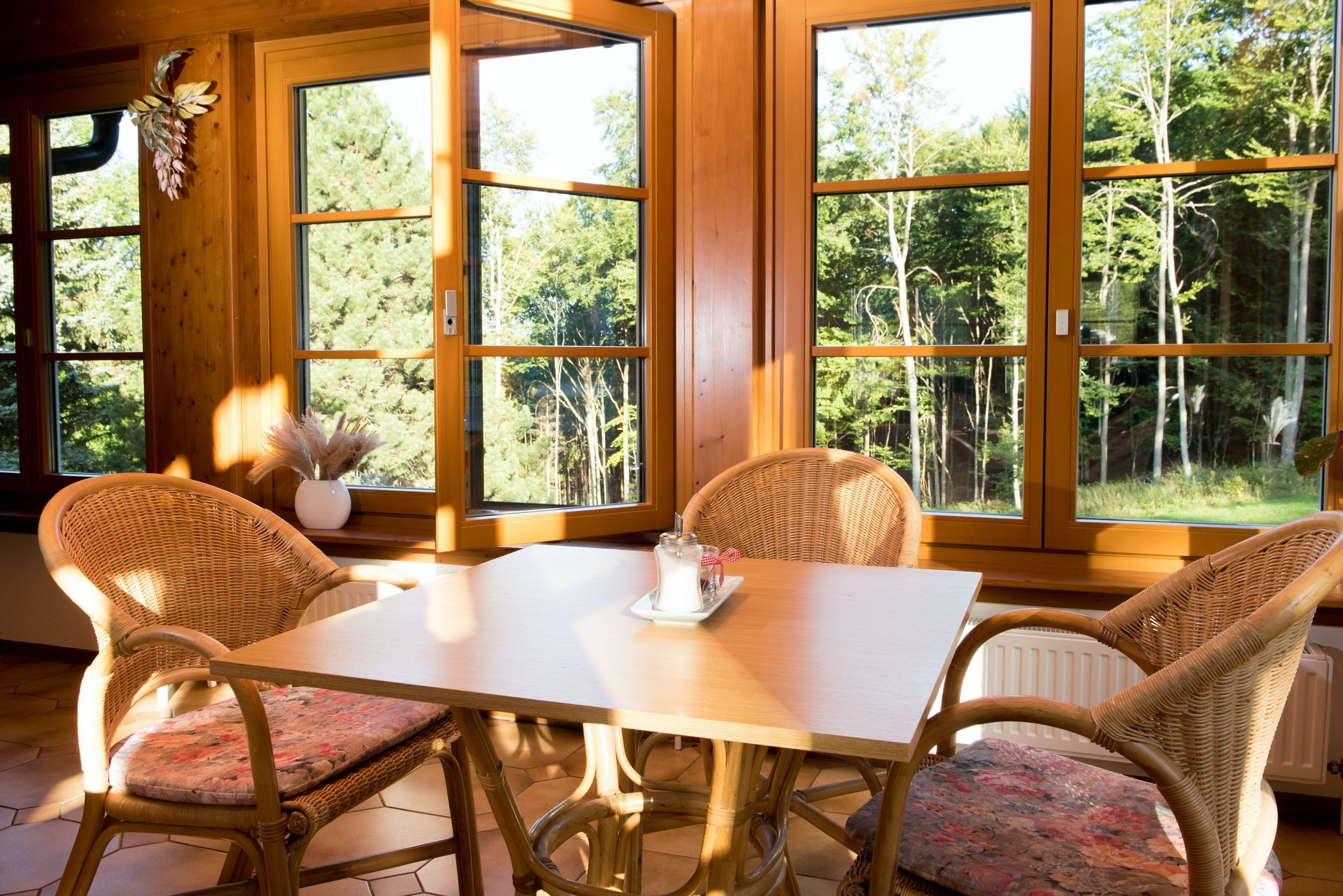 Ein Tisch mit vier Rattanstühlen in einem sonnigen Raum mit großen Fenstern und Blick auf einen Wald.
