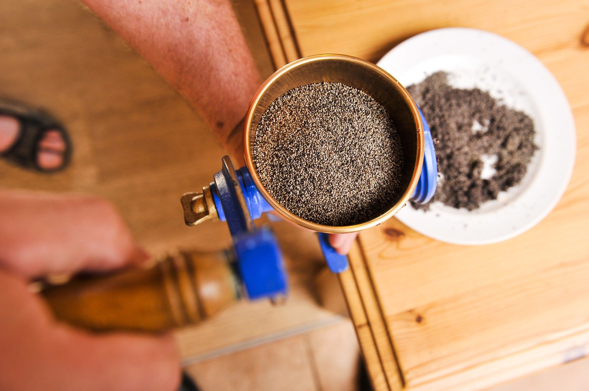 Nahaufnahme einer Hand, die eine Mühle mit Mohnsamen bedient, daneben ein Teller mit gemahlenem Mohn.