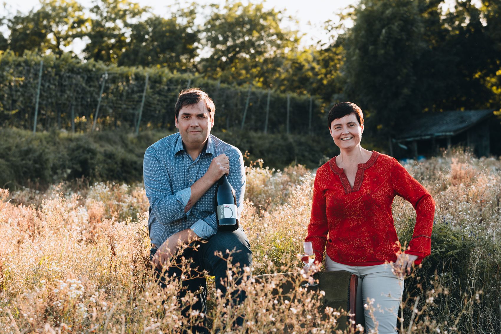 Zwei Personen in einem Weinberg, eine hält eine Weinflasche, die andere ein Weinglas.