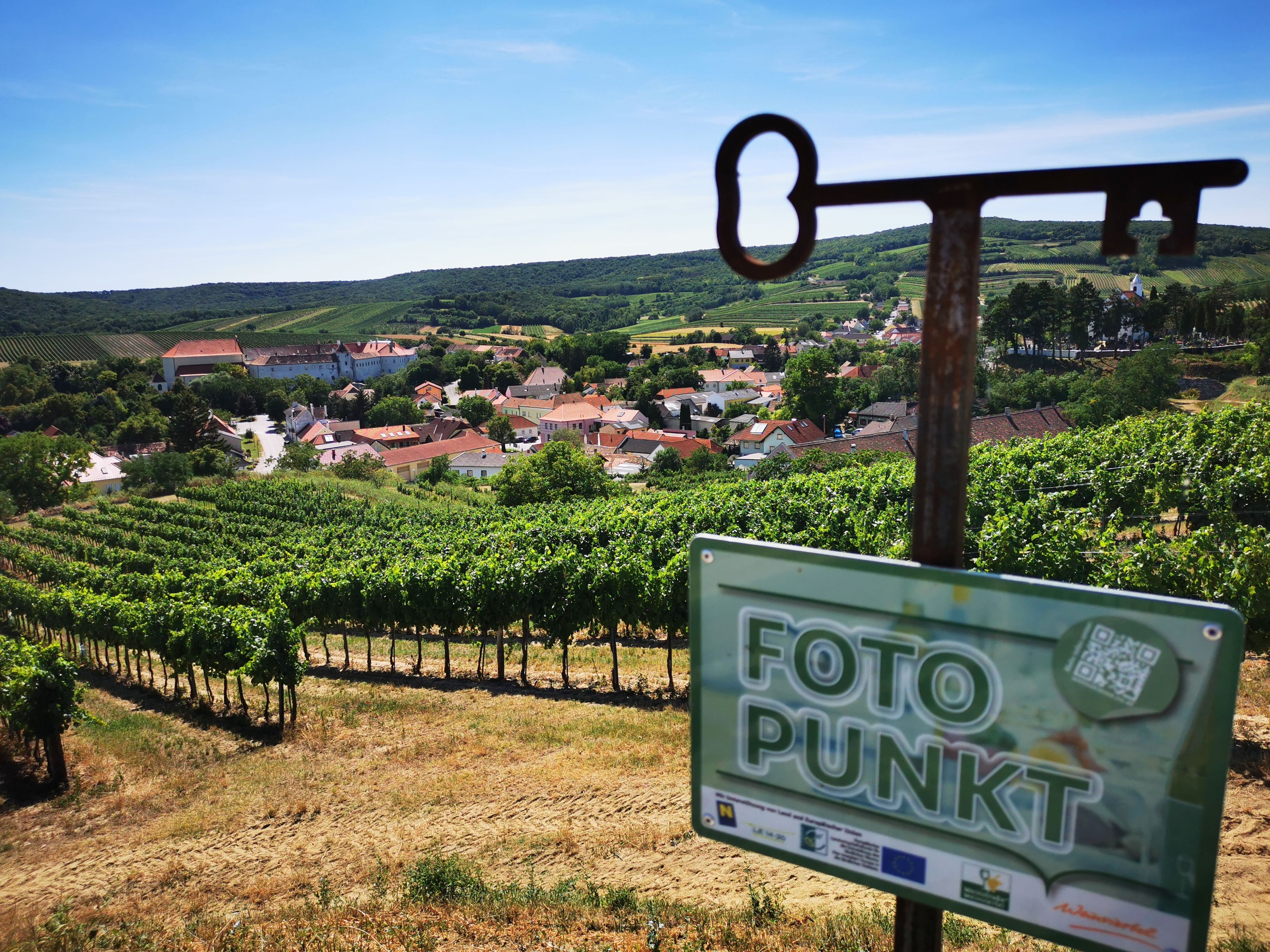 Blick auf ein Dorf inmitten von Weinbergen mit einem Schild 'Fotopunkt' im Vordergrund.