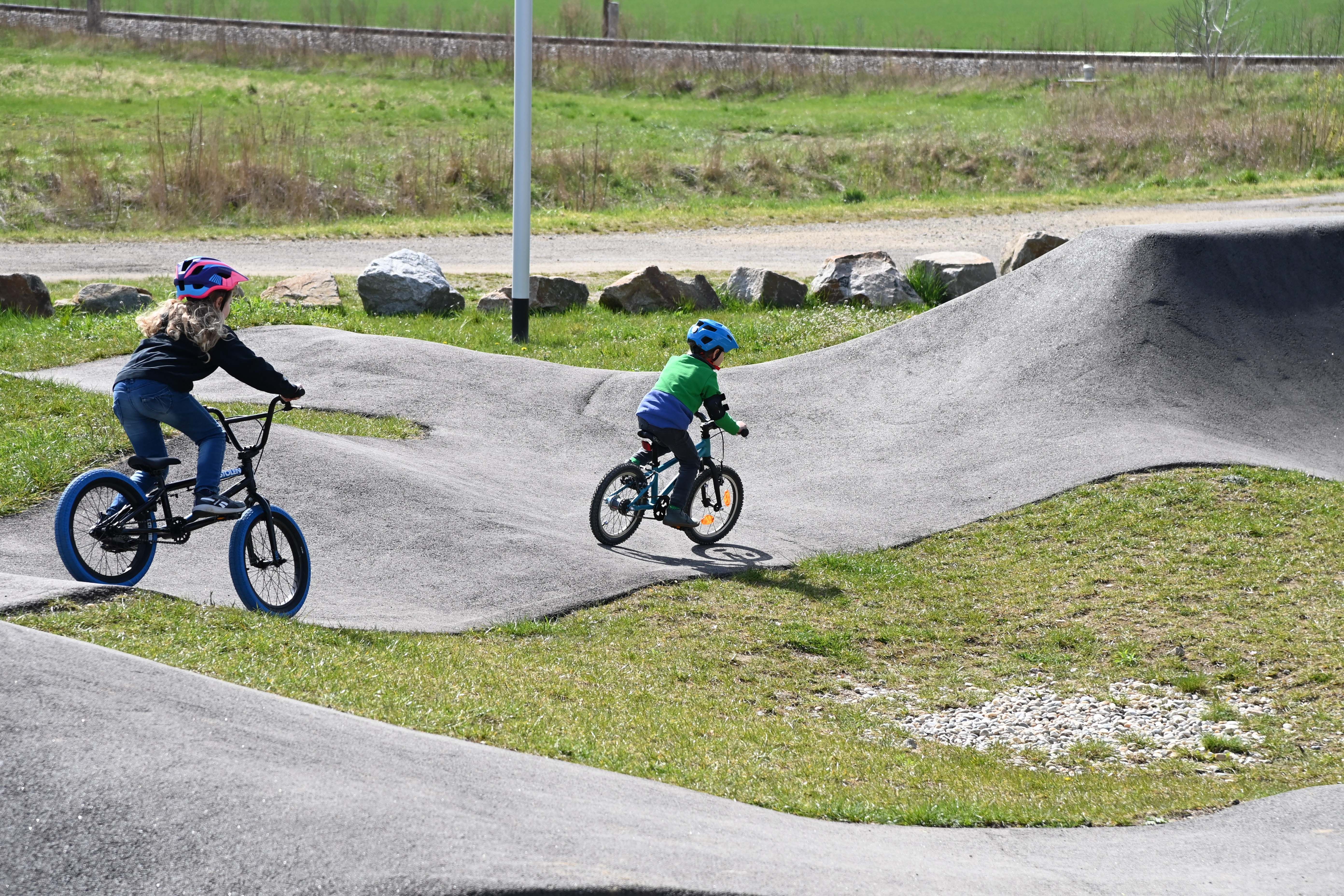 Zwei Kinder fahren mit Fahrrädern auf einem Pumptrack im Freien.