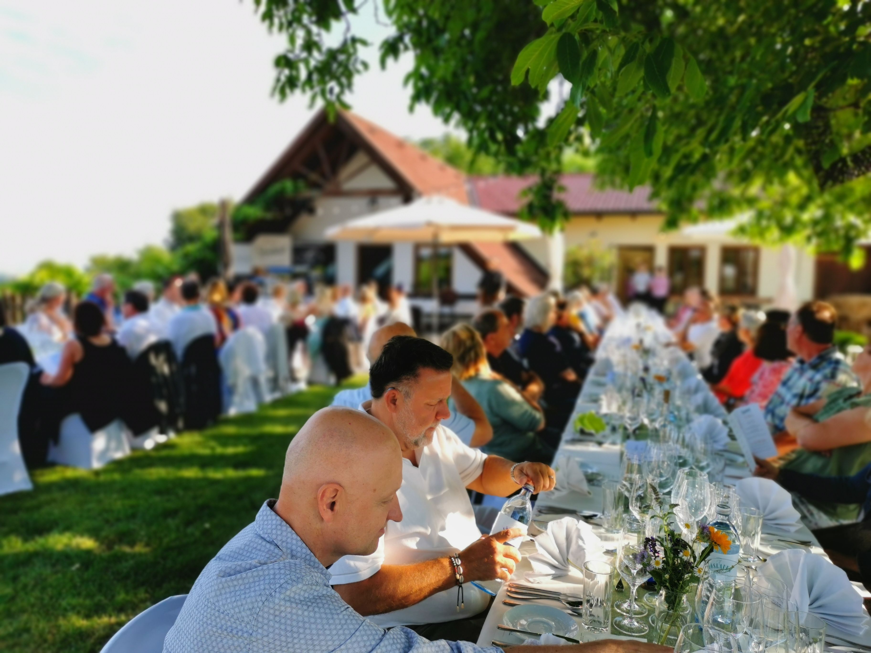 Menschen sitzen an langen Tischen im Freien bei einer Veranstaltung.