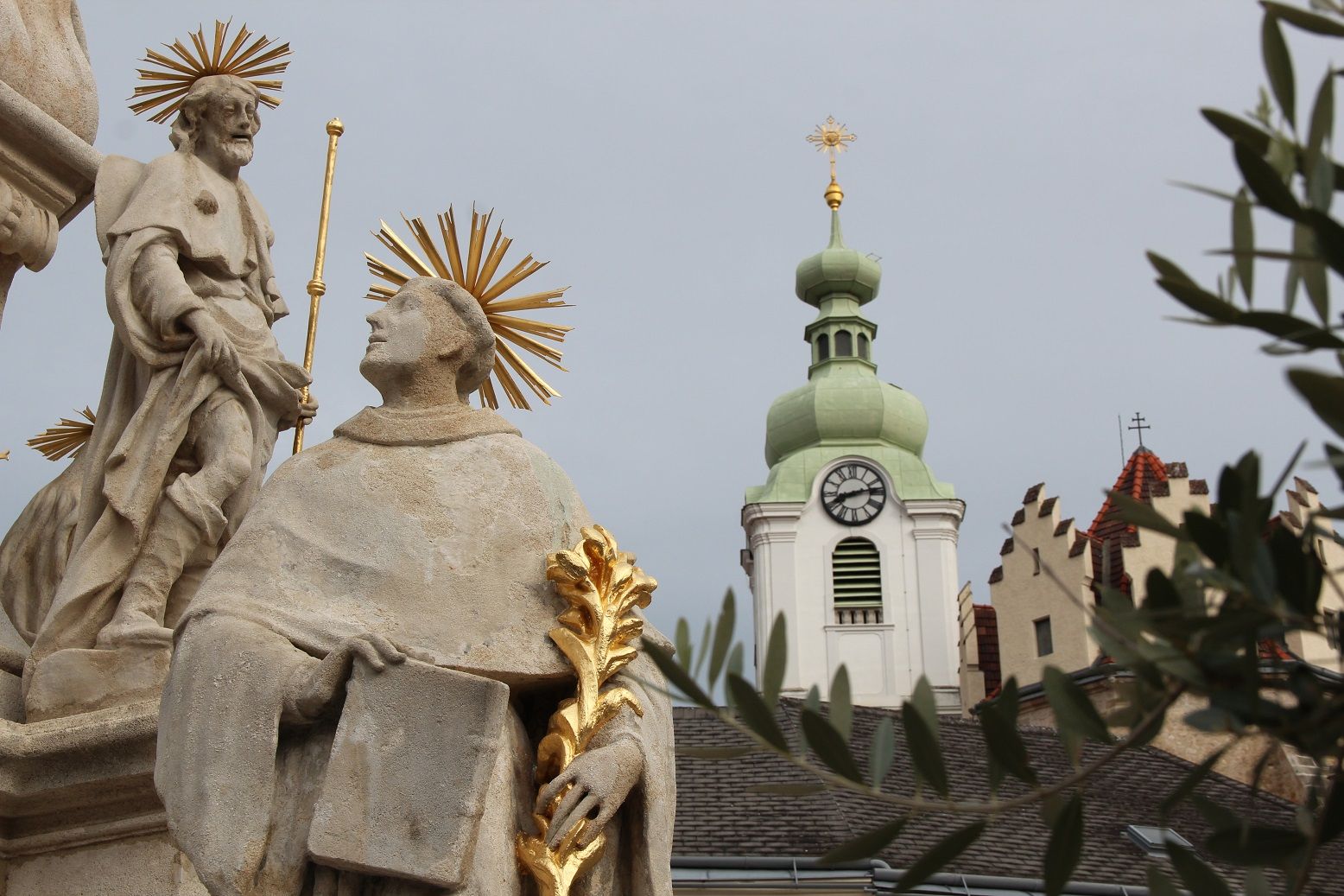 Statue mit Heiligenschein vor Kirchturm mit Uhr und grünem Dach.
