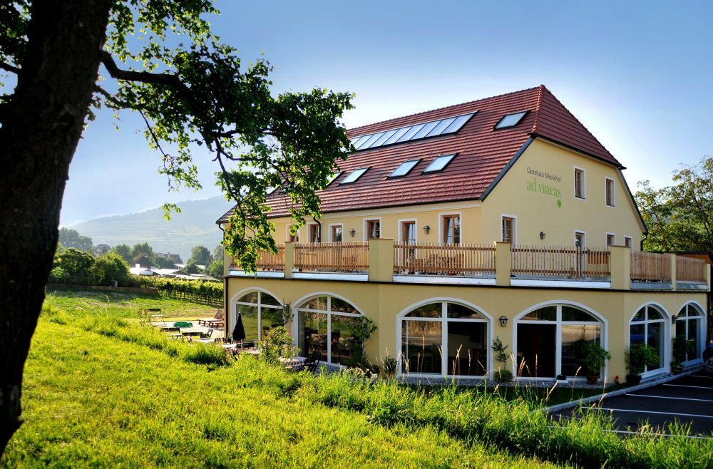 Gelbes Gebäude mit rotem Dach und Terrasse, umgeben von grüner Landschaft.