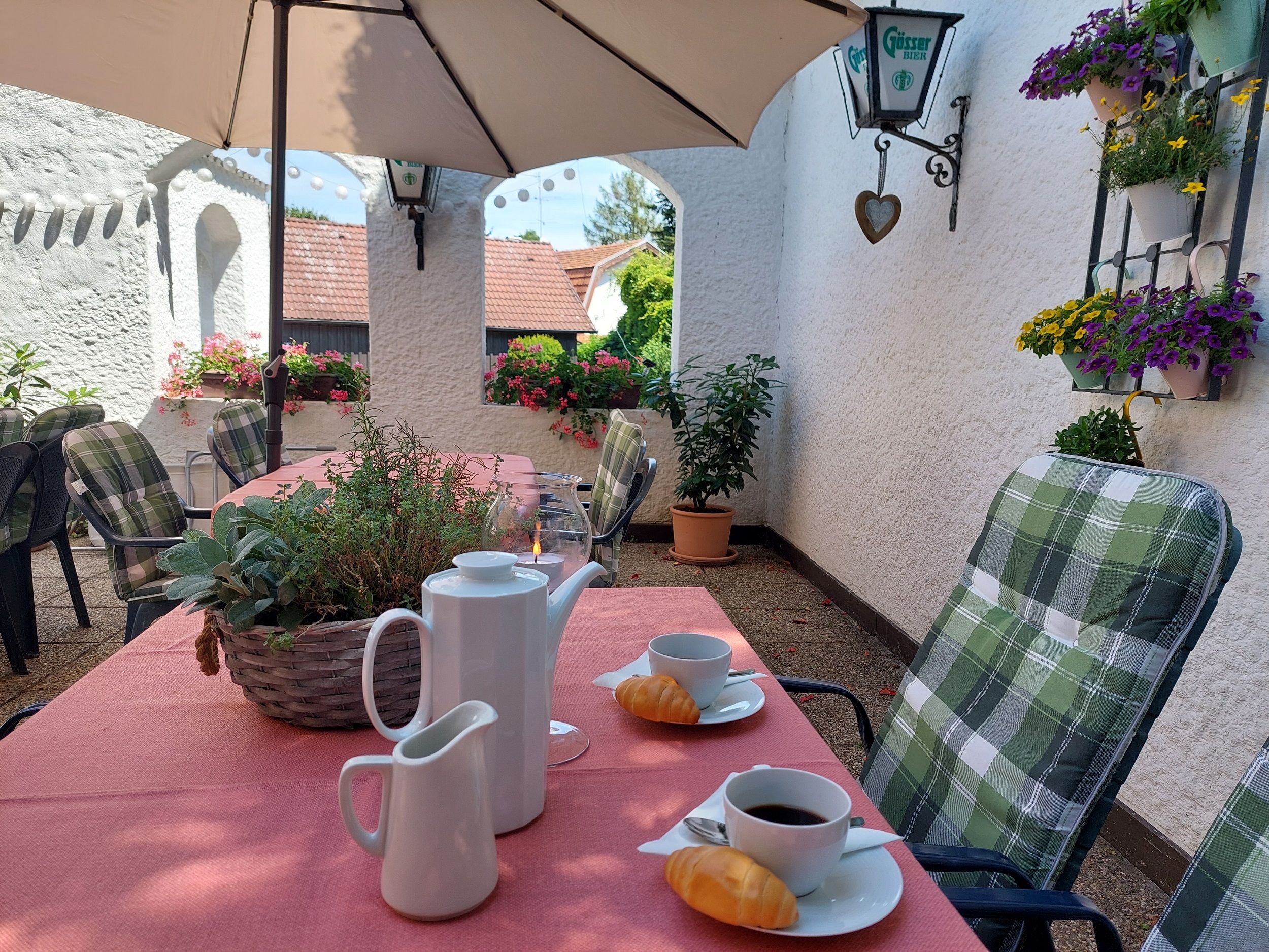 Gemütlicher Außenbereich mit gedecktem Tisch, Kaffeetassen und Gebäck, umgeben von Blumen und Pflanzen.