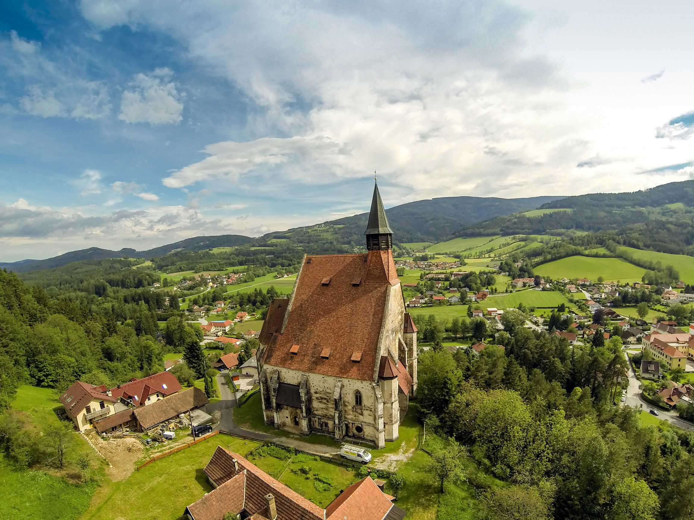 Luftaufnahme einer Kirche in einer ländlichen Umgebung mit Hügeln und Wäldern im Hintergrund.