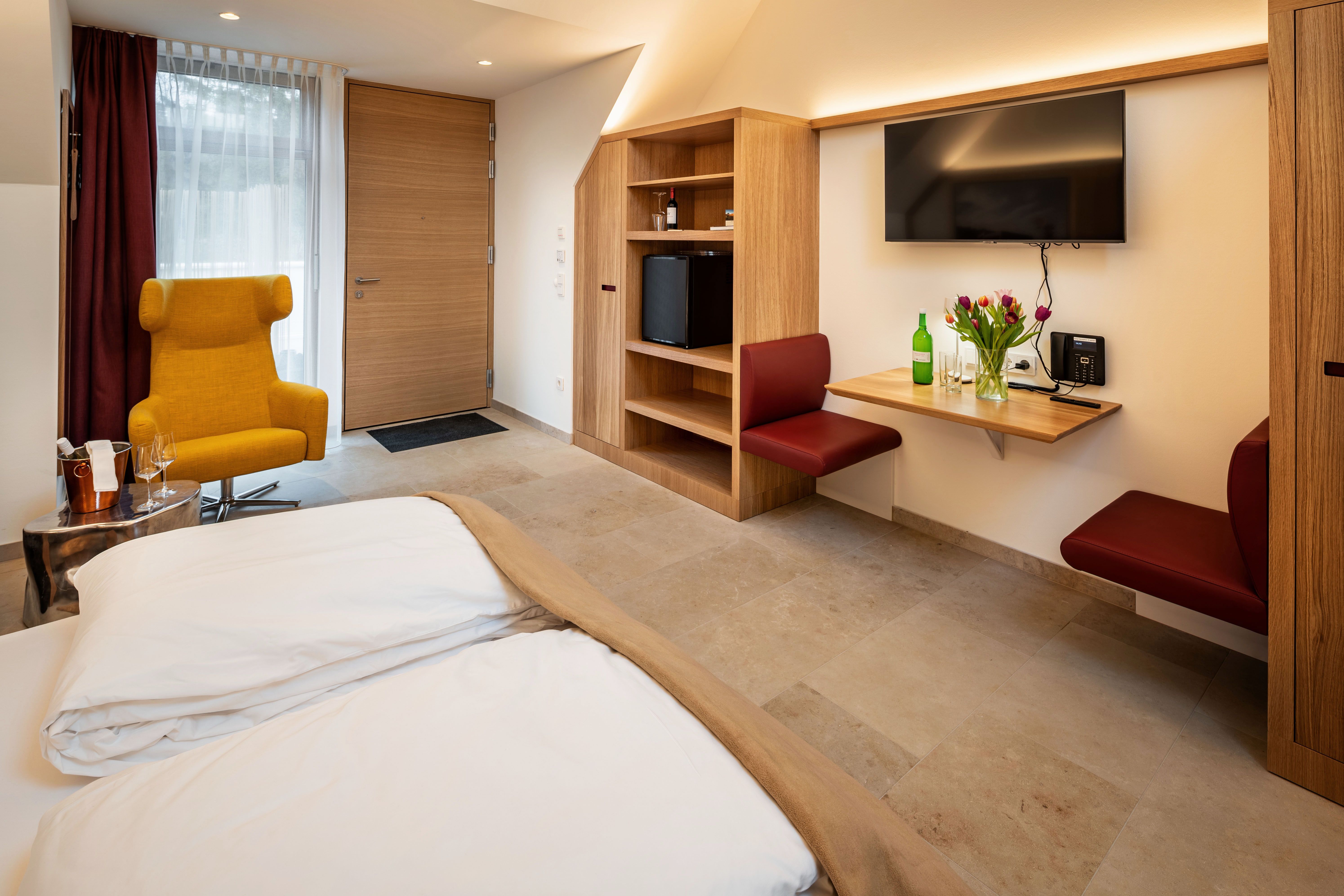Modern eingerichtetes Hotelzimmer mit gelbem Sessel, Holzmöbeln und Blumen auf einem Tisch.