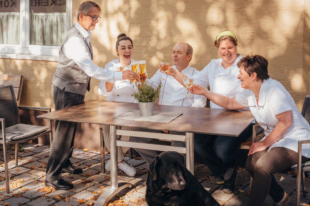 Ein Team von fünf Personen stößt draußen an einem Tisch an, mit einem schwarzen Hund im Vordergrund.