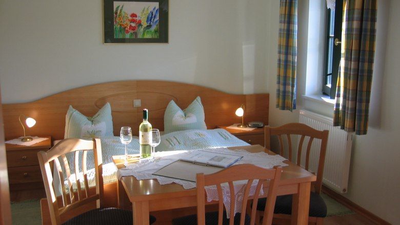Gemütliches Gästezimmer mit Doppelbett, Tisch und Stühlen, Weinflasche und Gläsern, Fenster mit Vorhängen.