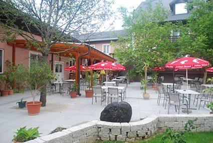 Ein Gastgarten mit Tischen, Stühlen und roten Sonnenschirmen auf einer Terrasse mit Bäumen und Pflanzen.