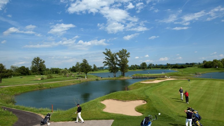 Golfplatz mit Wasserhindernissen und Spielern unter blauem Himmel.