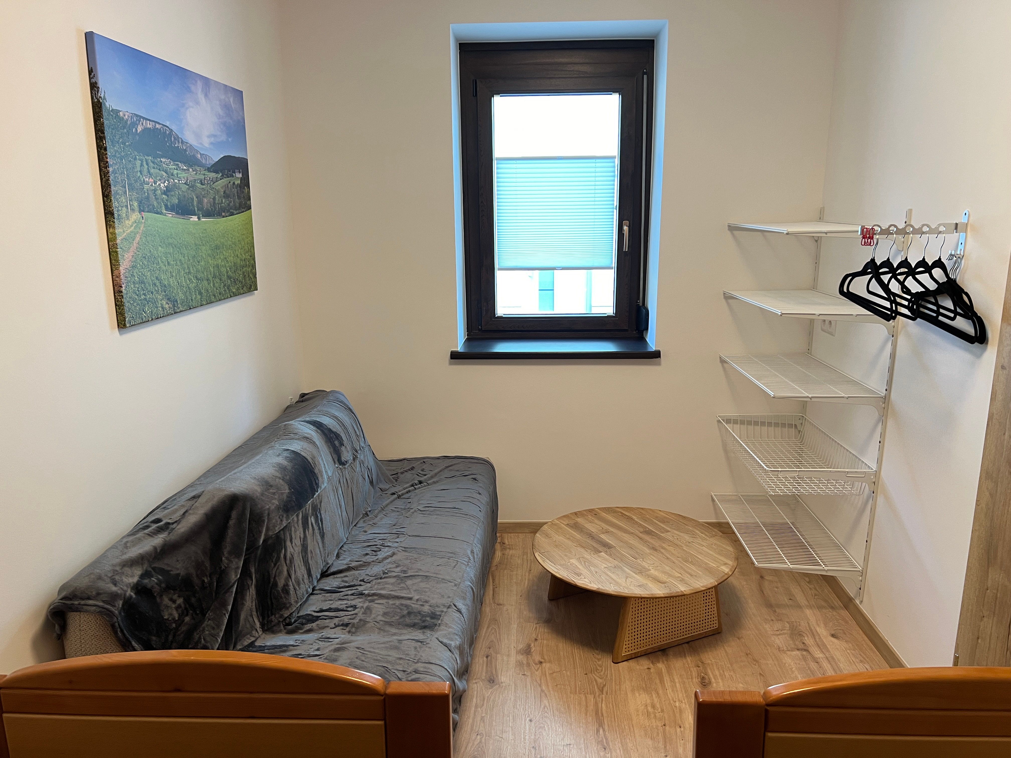 Kleines Zimmer mit Sofa, rundem Holztisch, Wandregal und Fenster.