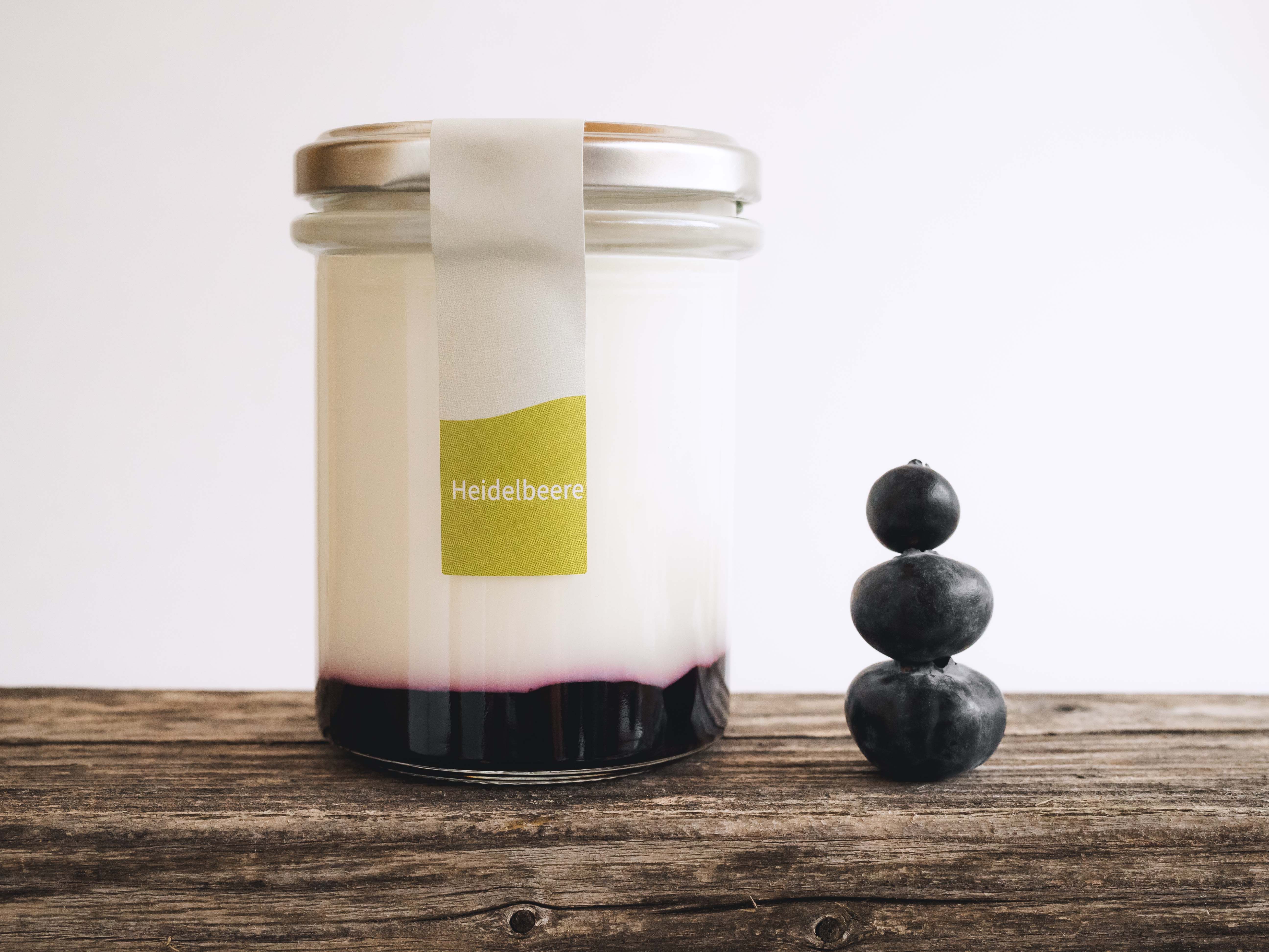Ein Glas gefüllt mit Joghurt und Fruchtspiegel auf einem Holztisch, daneben 3 gestapelte Heidelbeeren.