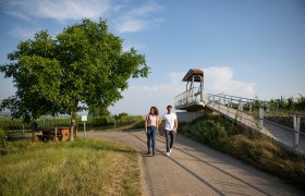Zwei Personen spazieren auf einem Weg neben einer Aussichtsplattform in einer l&auml;ndlichen Umgebung.