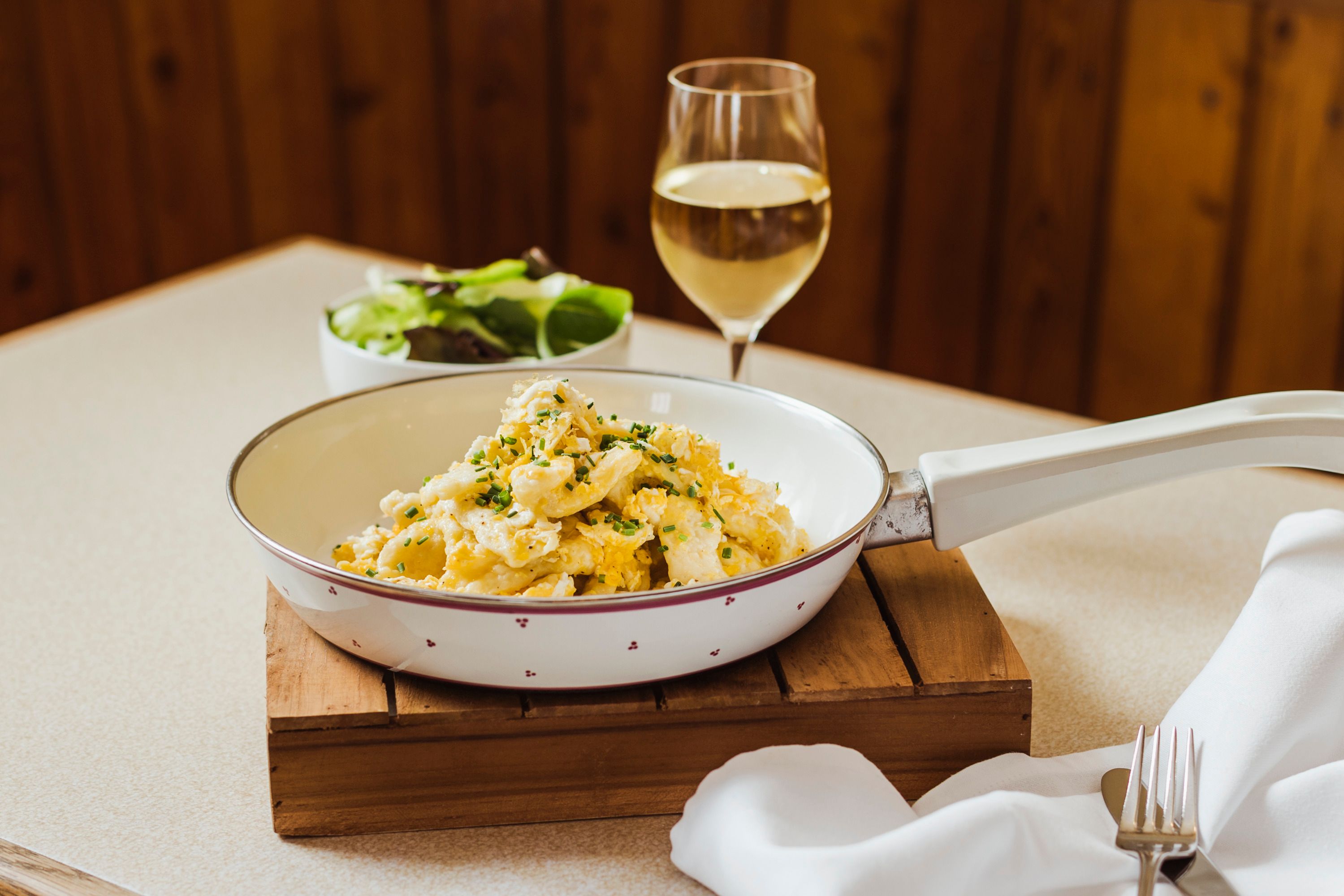Eiernockerl in einer Pfanne mit einem Glas Weißwein und einem Salat im Hintergrund.