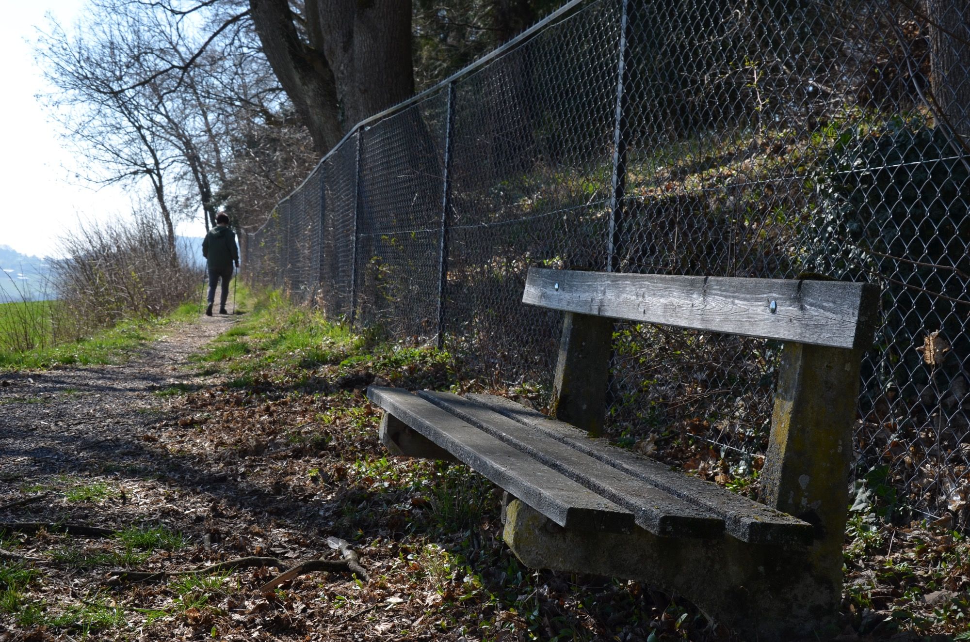 Ein Wanderweg mit einer Holzbank und einem Maschendrahtzaun. Eine Person geht in der Ferne.