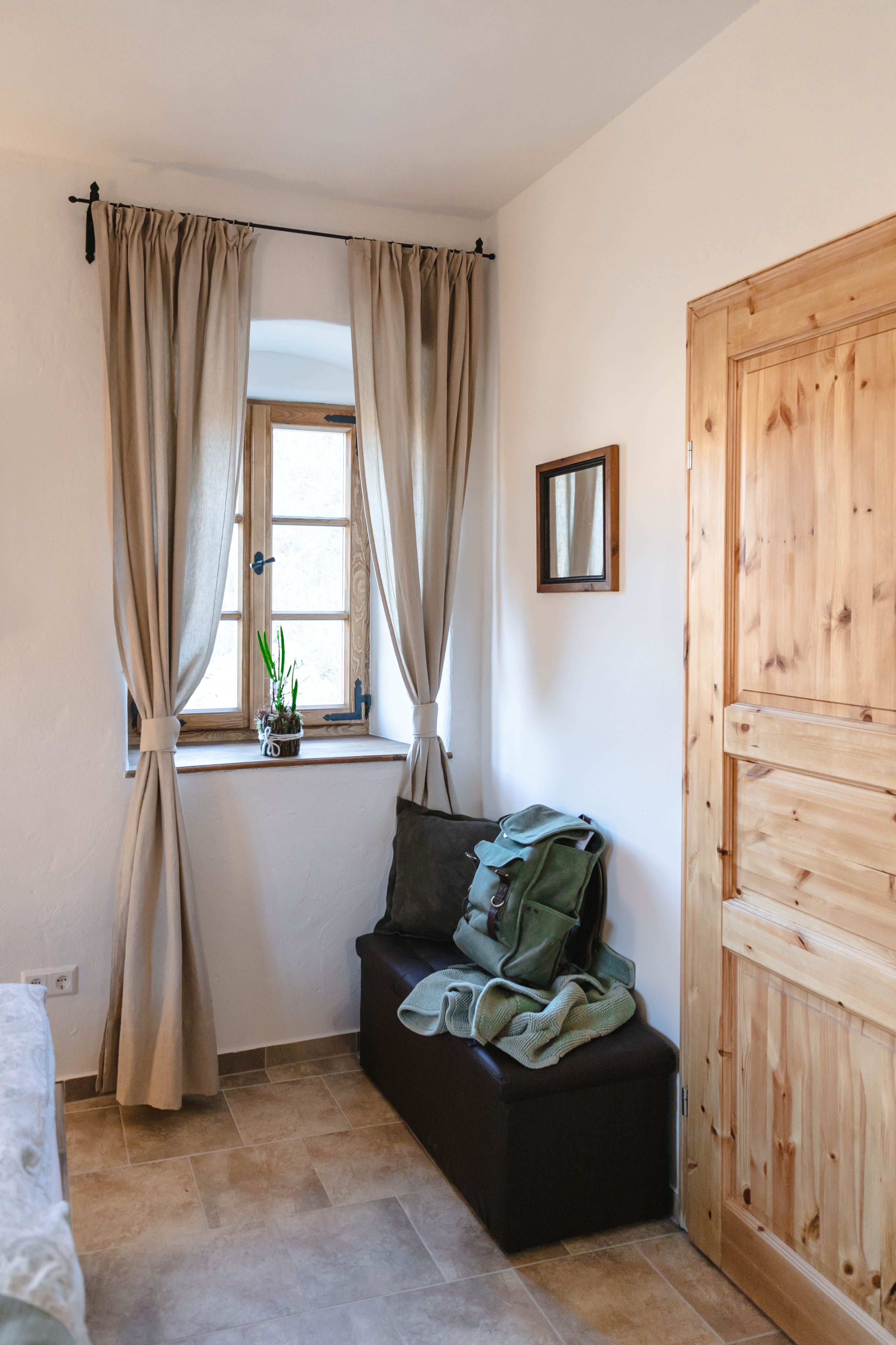 Ein kleiner Vorraum mit einem Fenster, Vorhängen, einer Holztür und einer Sitzbank mit Rucksack und Decke.