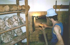 Ein B&auml;cker in einem blauen Tanktop und Hut nimmt Brot aus einem Ofen. Im Hintergrund sind Regale mit Brotlaiben zu sehen.