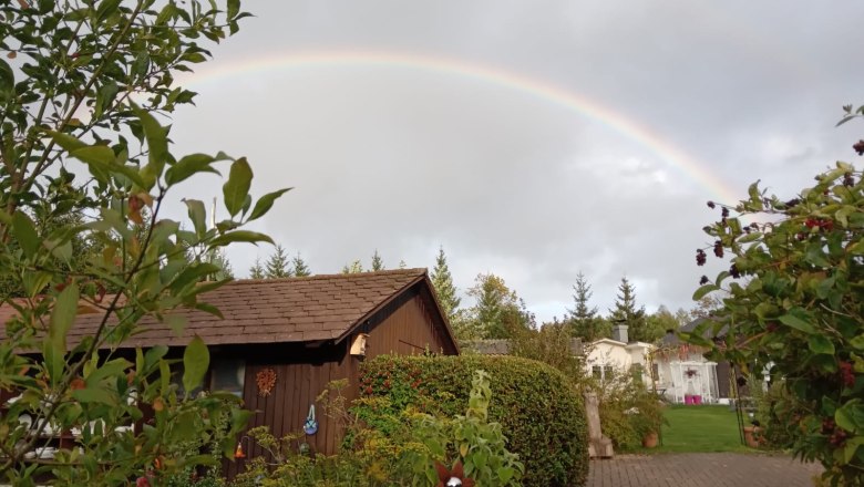 Ein Regenbogen spannt sich &uuml;ber einem Garten mit einem Holzh&auml;uschen und Pflanzen.