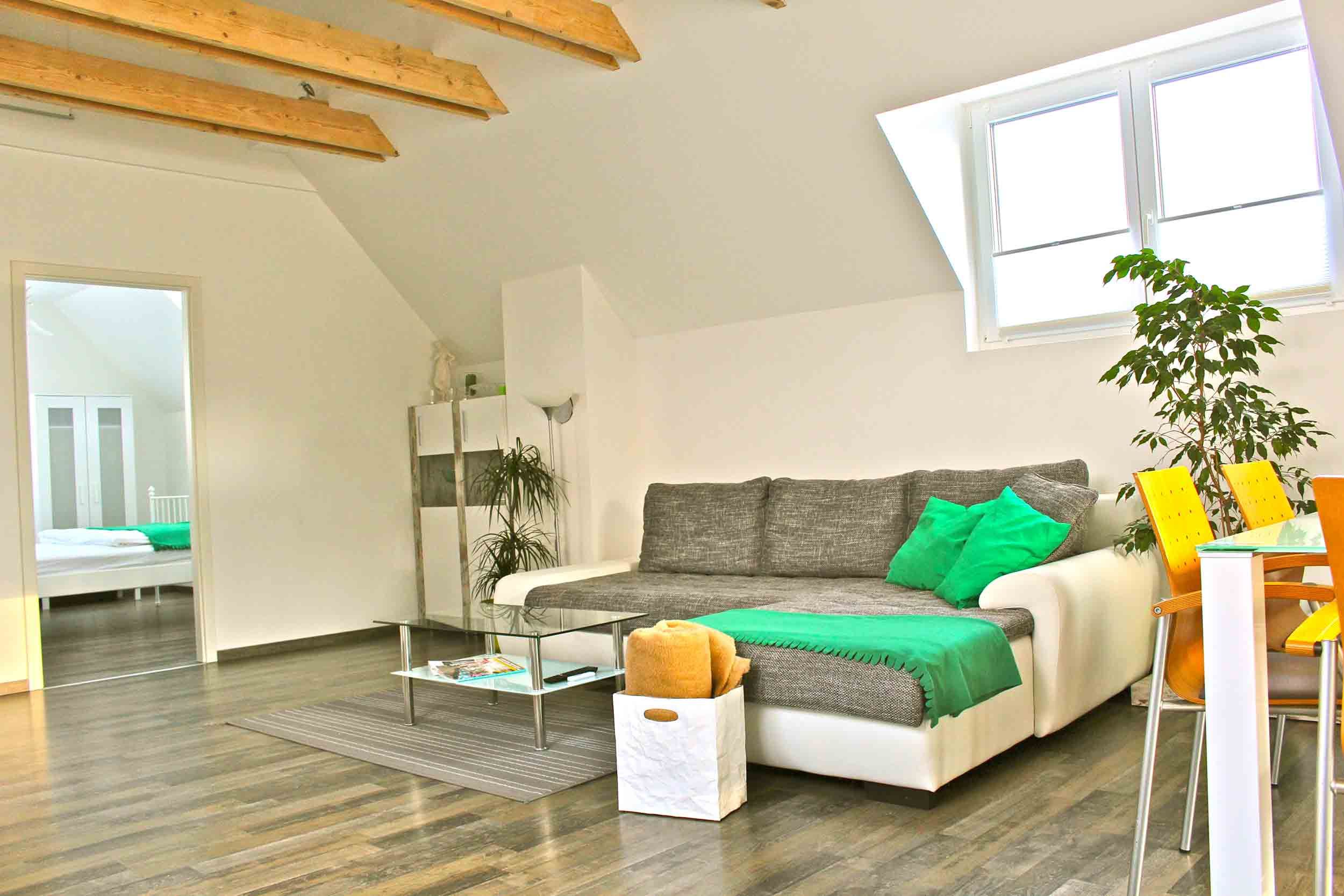 Helles Wohnzimmer mit grauem Sofa, grünen Kissen, Glastisch und Essbereich mit gelben Stühlen.