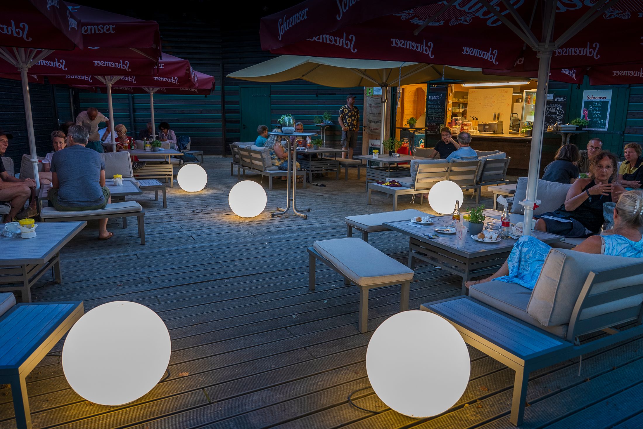 Menschen sitzen in einer gemütlichen Lounge am See mit leuchtenden Kugeln und Sonnenschirmen.