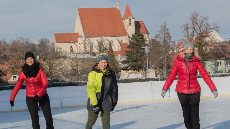 Drei Personen beim Eislaufen auf einer Eisbahn mit einer Kirche im Hintergrund.