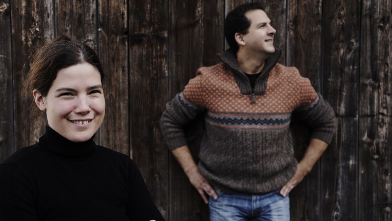 Zwei Personen vor einer Holzwand, eine Frau im Vordergrund l&auml;chelt, ein Mann im Hintergrund schaut zur Seite.