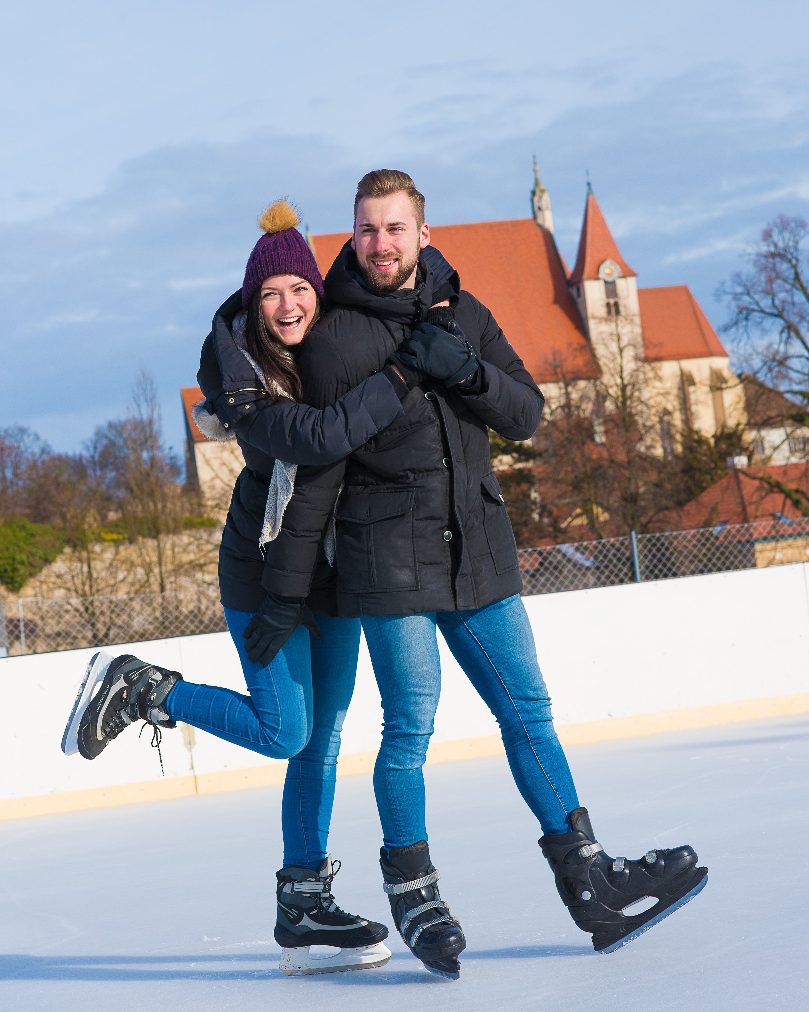 Ein Paar auf einer Eislaufbahn, im Hintergrund eine Kirche mit rotem Dach.
