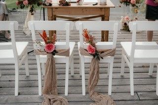 Weiße Stühle mit Blumen und Stoff dekoriert auf einer Holzterrasse, vorbereitet für eine Hochzeit.