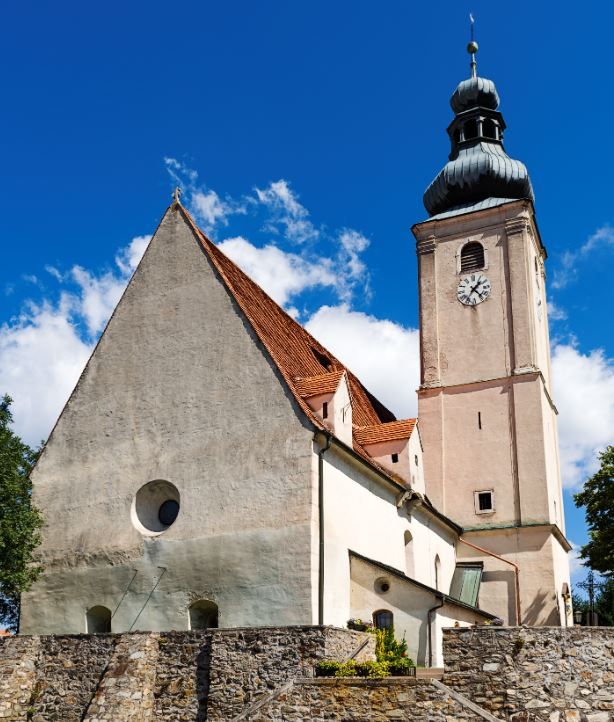 Pfarrkirche Wiesmath mit Turm, Friedhofsmauer aus Stein und Uhr vor blauem Himmel.