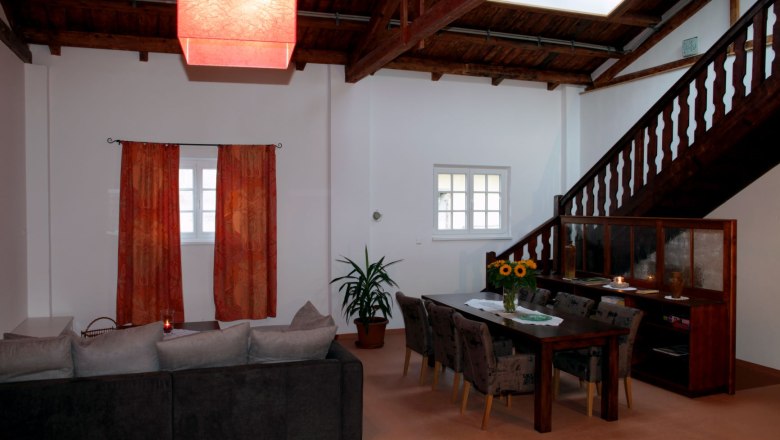 Wohn- und Essbereich in einem Loft mit Holzbalkendecke, rotem Lampenschirm, grauem Sofa, Esstisch mit St&uuml;hlen und Treppe.