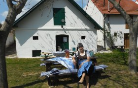 Eine Frau in traditioneller Kleidung sitzt auf einer Bank vor einem wei&szlig;en Geb&auml;ude mit gr&uuml;nen Fensterl&auml;den.