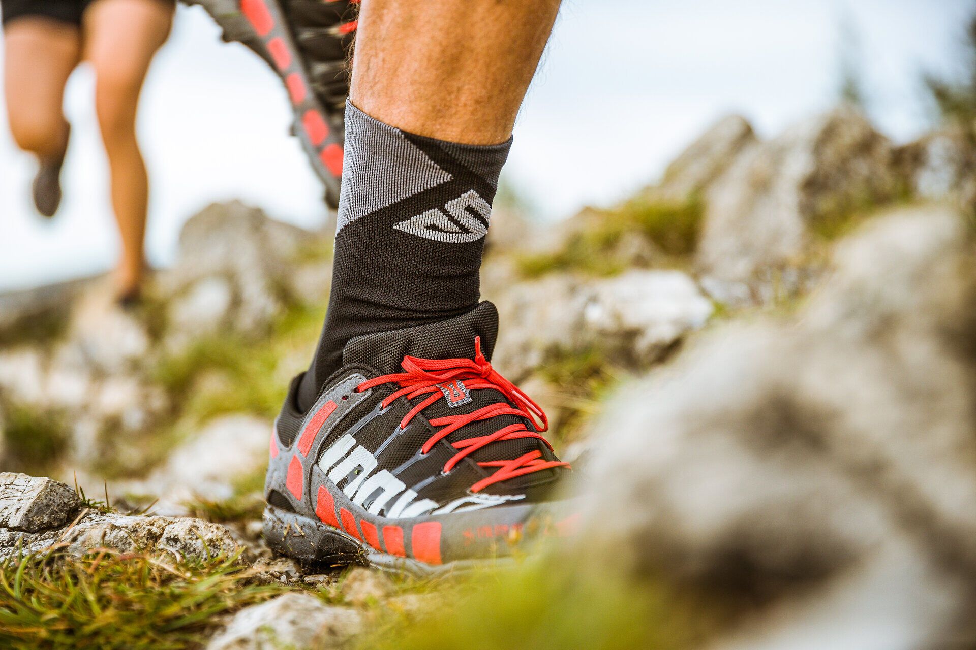 Ein Bein in einem Laufschuh in Detailaufnahme, die Person läuft auf einem natürlichem Untergrund mit Wiese und Steinen.