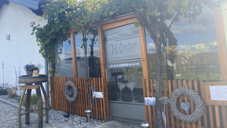 Eingang eines Geb&auml;udes mit Holzverkleidung und Weinreben, dekoriert mit Kr&auml;nzen und einem Tisch mit Blumen.