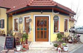 Eingang vom Bauernladen Stocker mit gelber Fassade, roten Dachziegeln und Pflanzen und Deko im Vordergrund.