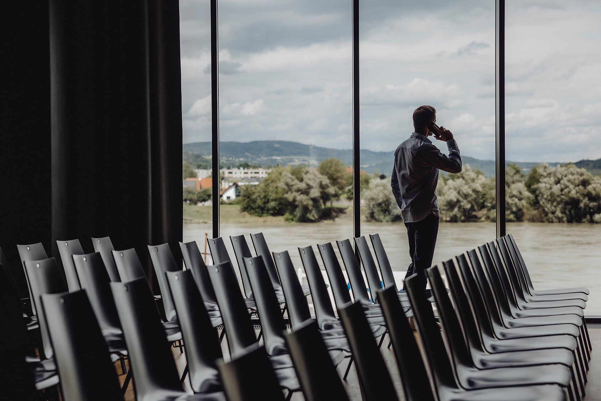 Die Atmosphäre in der Stadthalle Ybbs ist geprägt von einer harmonischen Verbindung zwischen modernem Design und der atemberaubenden Natur der Donau. Die großen Fenster bieten einen malerischen Blick auf die sanften Hügel und das glitzernde Wasser, während die leeren Stühle auf die bevorstehenden Begegnungen und Erlebnisse warten.