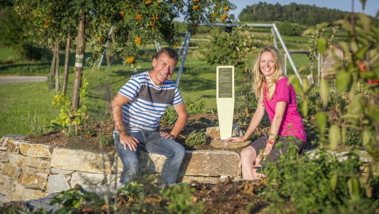 Zwei Personen sitzen auf einer Steinmauer in einem Garten mit Pflanzen und einem Schild.