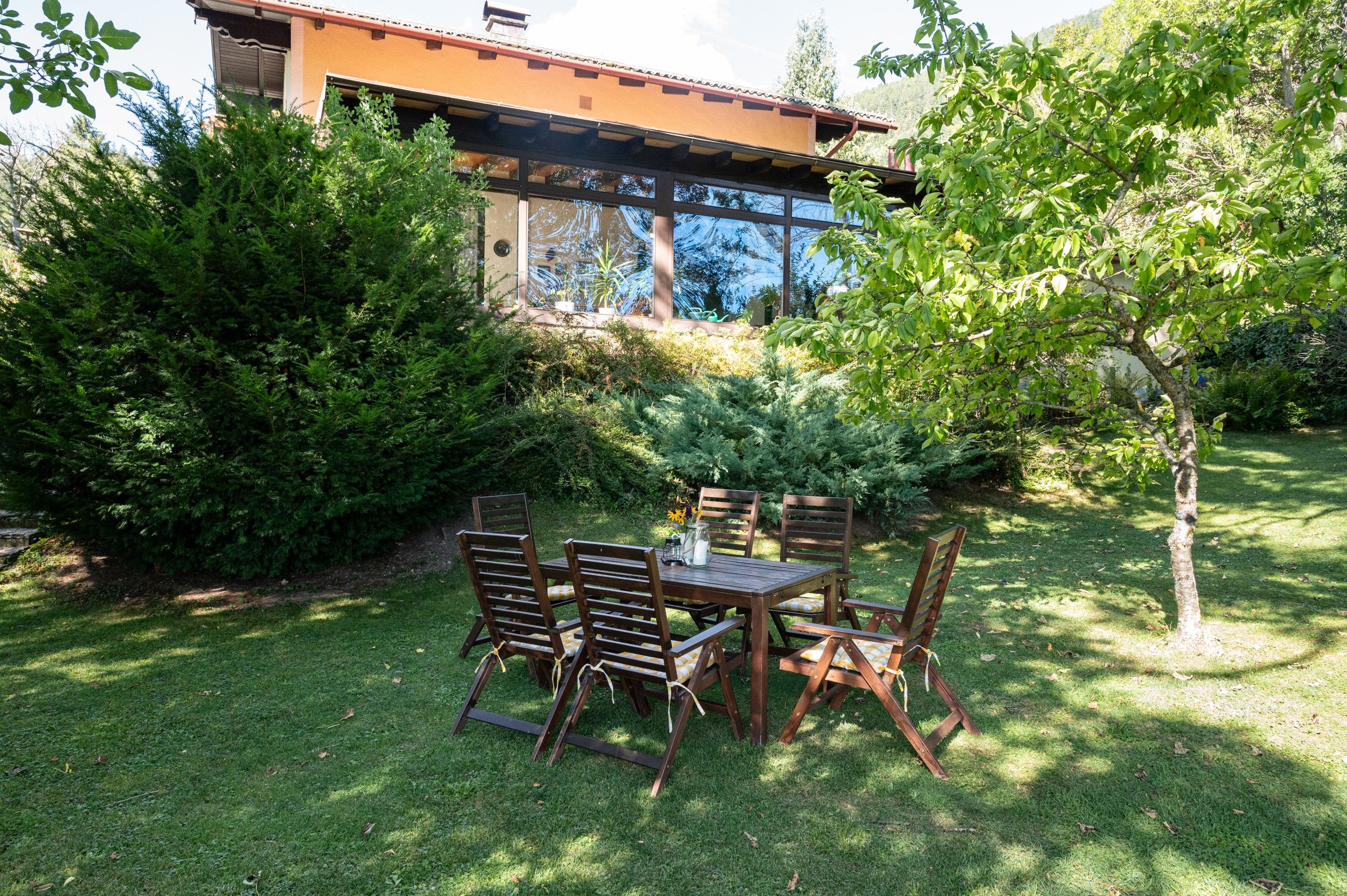 Ein Garten mit Holztisch und Stühlen vor einem Ferienhaus mit großen Fenstern und umgeben von Bäumen und Sträuchern.