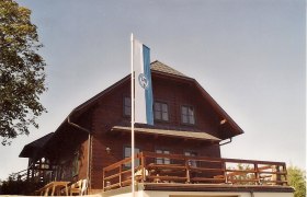 Holzhütte mit Fahne auf Terrasse, blauer Himmel.