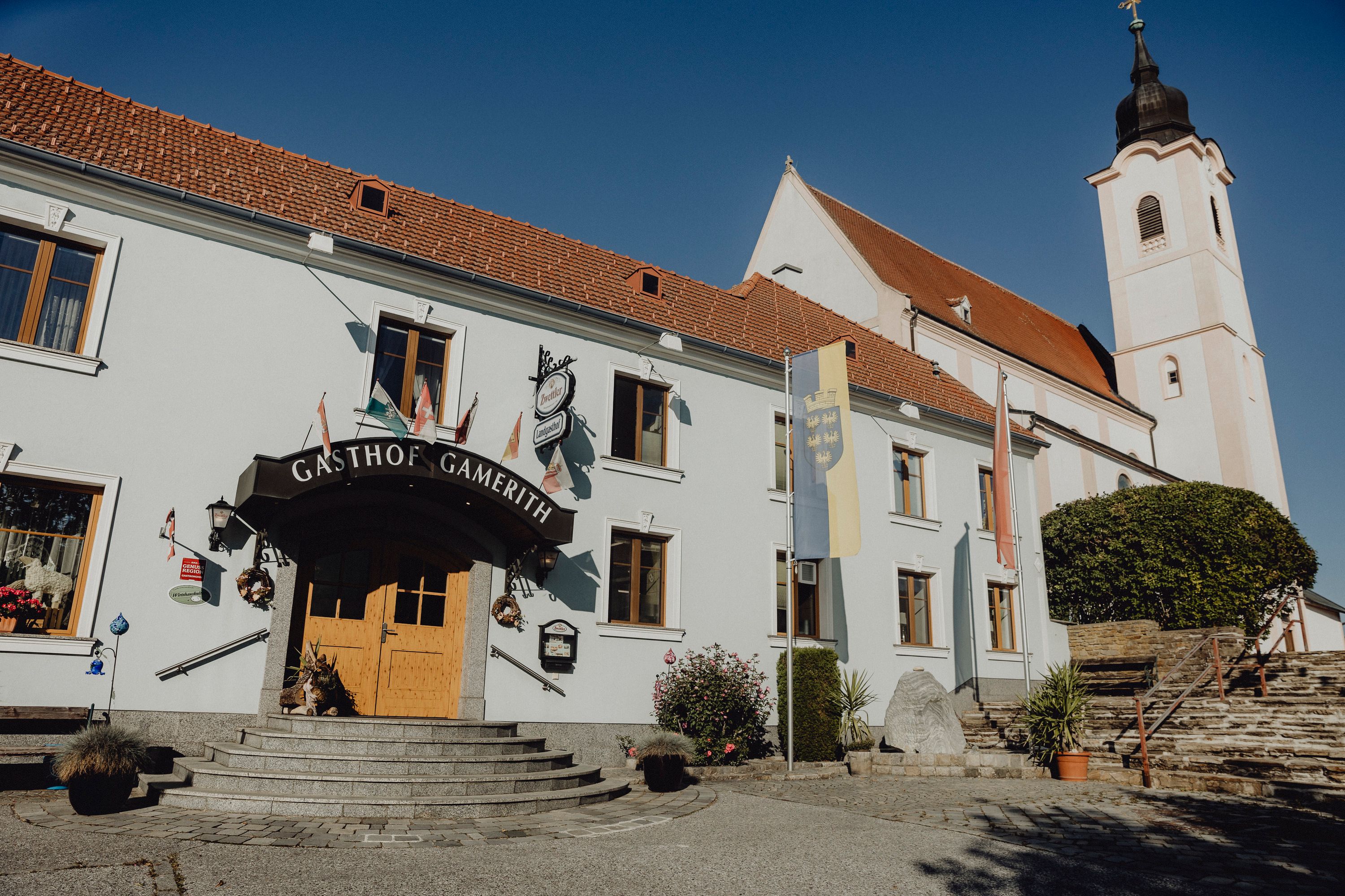 Ein Gasthof mit der Aufschrift 'Gasthof Gamerith' neben einer Kirche mit Turm unter blauem Himmel.